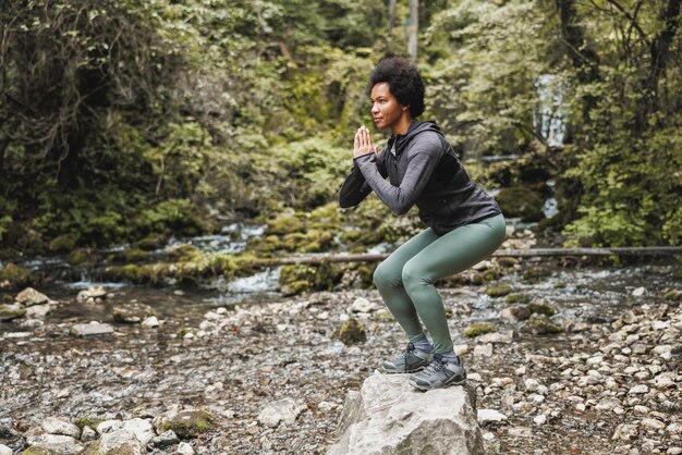 Belle coureuse afro-américaine mature faisant des exercices de squat après avoir fait du jogging près de la rivière dans la nature.