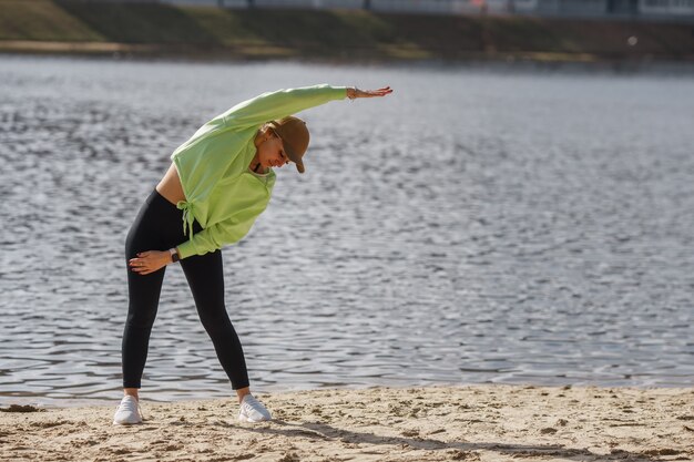 Belle coureuse autodéterminée en vêtements de sport à la mode posant isolée sur la rive du fleuve, étirement des jambes, échauffement des muscles avant de courir à l'extérieur tôt le matin