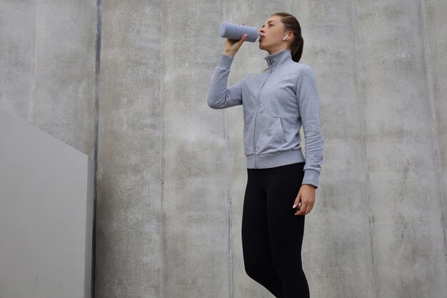 Belle coureuse debout à l'extérieur, buvant de l'eau de la bouteille. La femme de remise en forme fait une pause après avoir couru l'entraînement.