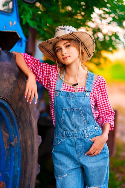 Belle cow-girl blonde en chapeau de paille et denim debout dans l'ensemble près d'un tracteur bleu.