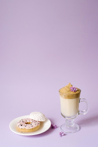 La belle dalgona boit un café mousseux dans une tasse transparente et des fleurs de lilas sur fond violet. Bonbons à l'assiette : Donat et guimauves tendres. Espace de copie