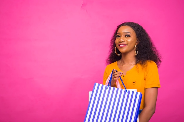 belle dame africaine isolée sur fond rose se sentant excitée alors qu'elle tient un sac à provisions.