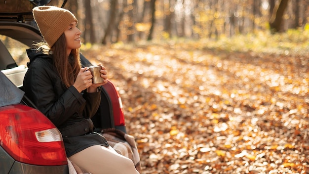 Photo belle dame assise dans un coffre de voiture ouvert et buvant du thé ou du café nature d'automne sur fond de feuilles d'érable colorées voyage d'aventure style de vie du voyageur