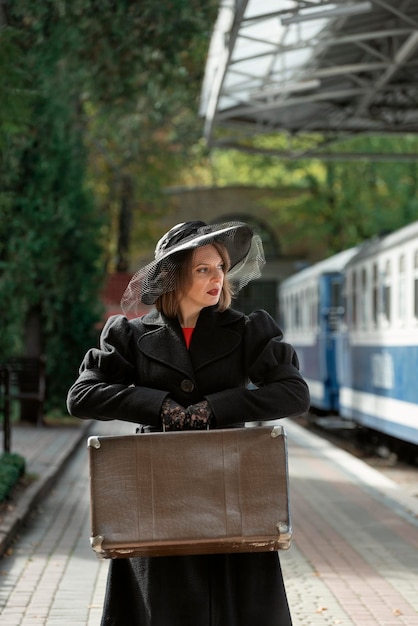 Photo belle dame au chapeau noir à larges bords avec voile tient une valise en cuir