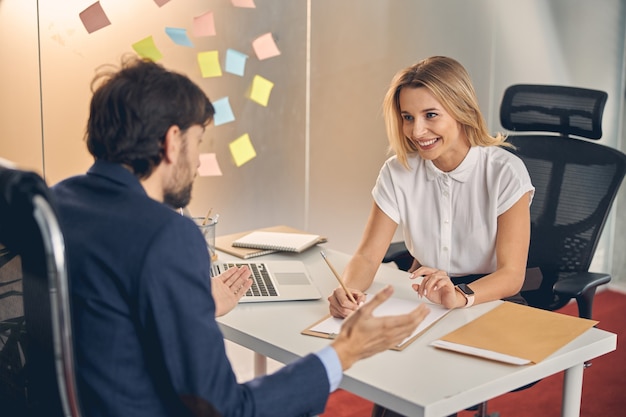 Belle dame blonde regardant un collègue et souriant tout en étant assise à la table avec un ordinateur portable et des documents