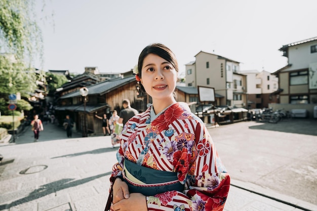 belle dame en robe kimono fleurie regarde vers le ciel bleu profiter du coucher du soleil sur la rue. touristes en arrière-plan visitant la vieille ville japonaise de kyoto au japon. jeune femme asiatique en tissu traditionnel souriant.