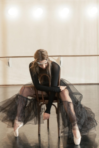 Photo belle danseuse de ballet contemporaine en vêtements noirs posant sur une chaise en bois sur une répétition dans un studio de danse