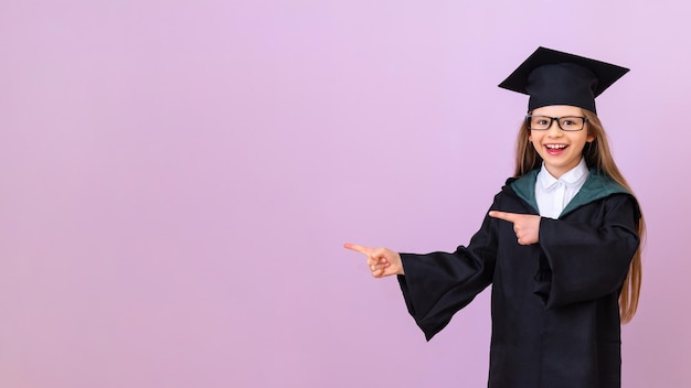 Une belle diplômée dans un bonnet de graduation et une robe de cérémonie pointe ses doigts vers une publicité sur un fond violet isolé. espace de copie. une écolière joyeuse. obtenir un certificat à l'école