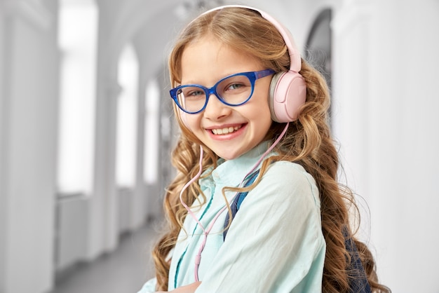 Belle écolière aux longs cheveux blonds portant une chemise bleue et des lunettes, écoutant de la musique avec des écouteurs