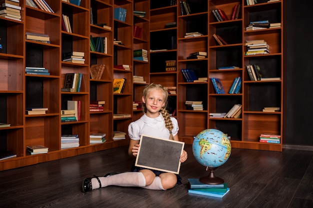 Une belle écolière blonde en uniforme est assise avec une ardoise et un globe dans la classe de l'école