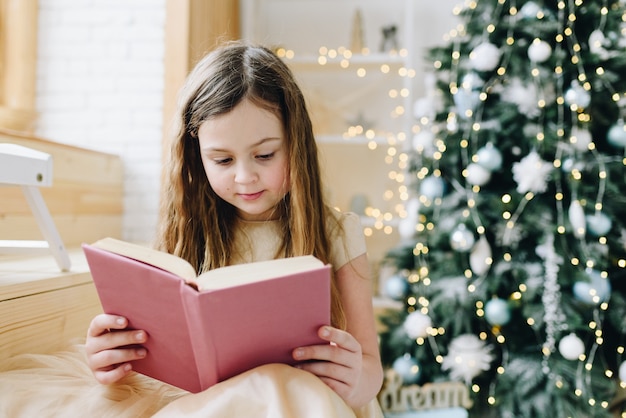 Belle écolière caucasienne lisant un livre violet près de l'arbre de Noël décoré