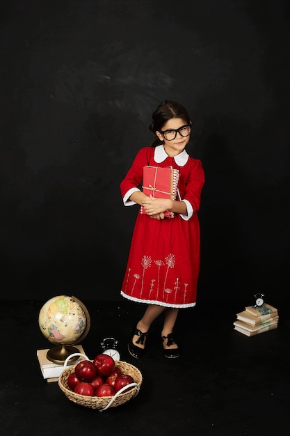 Une belle écolière dans une robe rouge avec un livre de pommes et globe sur un fond noir
