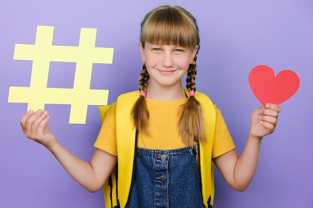 Photo belle écolière souriante attirante tenant un petit coeur rouge et un grand symbole de hashtag, heureuse de regarder la caméra, porte un sac à dos jaune, isolé sur fond violet en studio. retour au concept de l'école