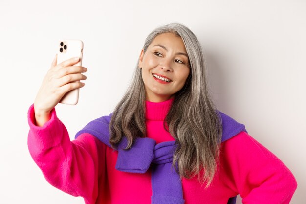 Belle et élégante femme senior asiatique prenant selfie sur smartphone, souriante et posant dans un pull rose à la mode, debout sur fond blanc