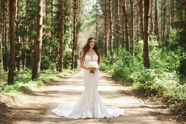 Belle et élégante fille modèle brune avec une coiffure décorée de bijoux en robe de dentelle à la mode posant dans la forêt tôt le matin