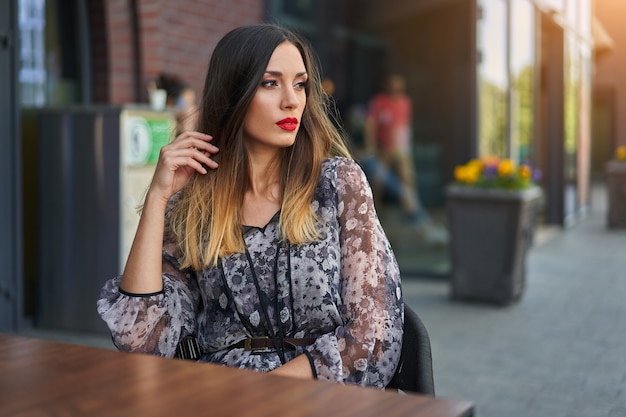 Belle élégante jeune fille aux cheveux longs teints en noir se trouve à un café en plein air de table d'été en attente de son petit ami.