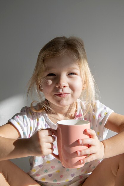 Belle enfant fille blonde tient une tasse rose sur le fond d'un mur blanc