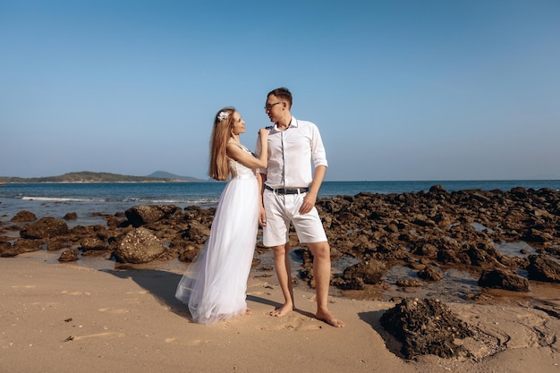 Belle épouse en robe de mariée blanche tenant affectueusement l'épaule de son mari bien-aimé en costume blanc sur fond de plage de galets et de mer. Notion d'amour