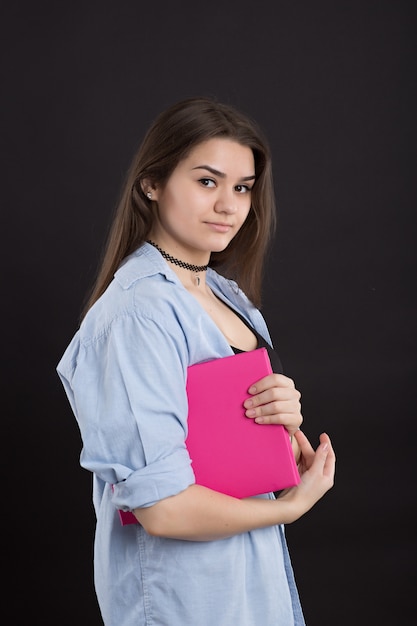 Belle étudiante en chemise en jean aux cheveux longs, sur mur noir, tient en mains un grand livre