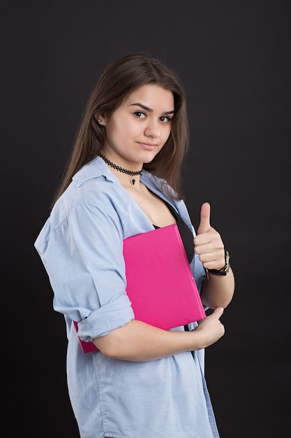 Belle étudiante en chemise en jean aux cheveux longs, sur mur noir, tient en mains un grand livre