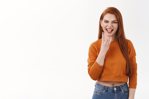 Une belle étudiante rousse impertinente et audacieuse fait la promotion de quelque chose d'excitant, d'activité ou d'événement. Jolie femme au gingembre montrant un geste rock-n-roll et une langue de bâton, mur blanc debout