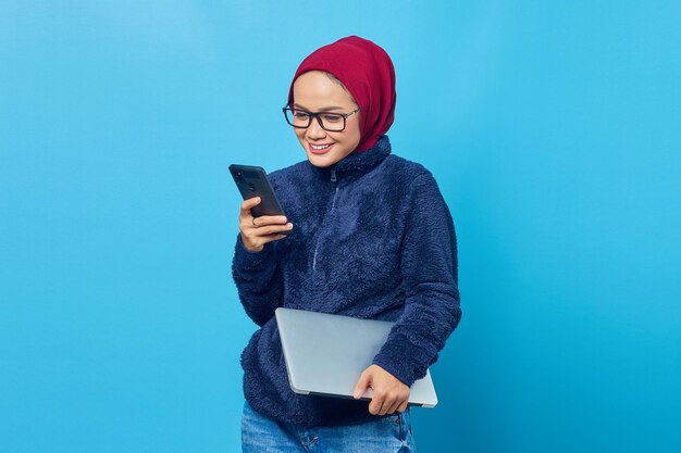 Belle étudiante souriante tenant un ordinateur portable et regardant un téléphone portable sur fond bleu