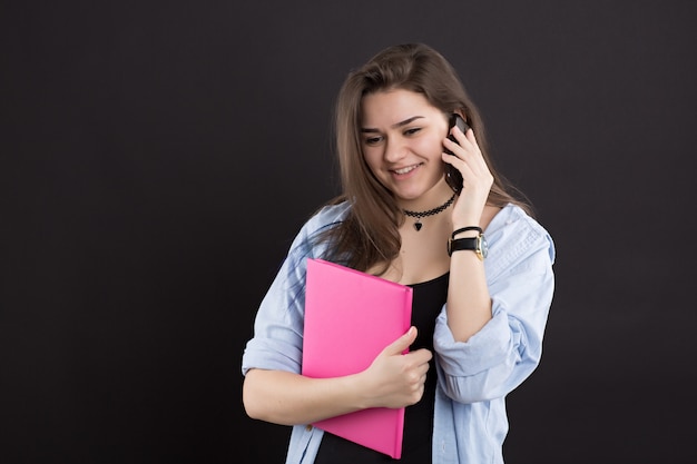 Belle étudiante surprise en chemise en jean aux cheveux longs, parlant au téléphone et tenant un livre