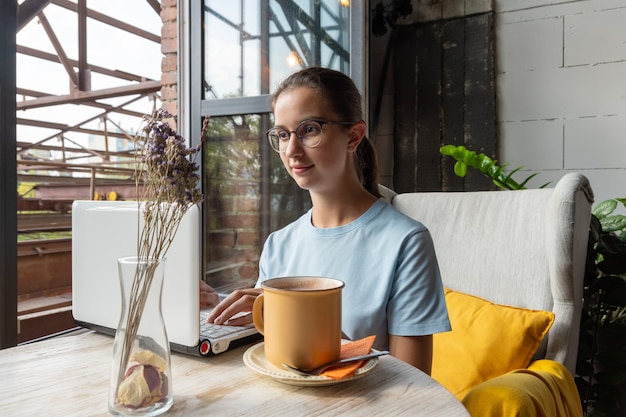 Belle étudiante utilisant un ordinateur portable, tapant un message, cherchant sur Internet assise à une table dans un café loft. Notion d'indépendant. Petit déjeuner du matin dans un café