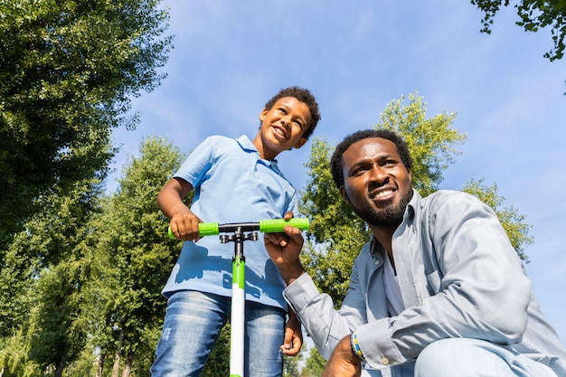 Belle famille afro-américaine heureuse au parc - Famille noire s'amusant à l'extérieur, papa enseignant à son fils à monter en scooter
