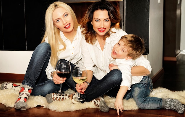 Belle famille composée d'une mère aux cheveux roux, d'une fille aux cheveux blonds et d'un fils assis sur le tapis de fourrure devant la cheminée. Le feu dans la cheminée brûle. Femmes tenant des verres de vin.