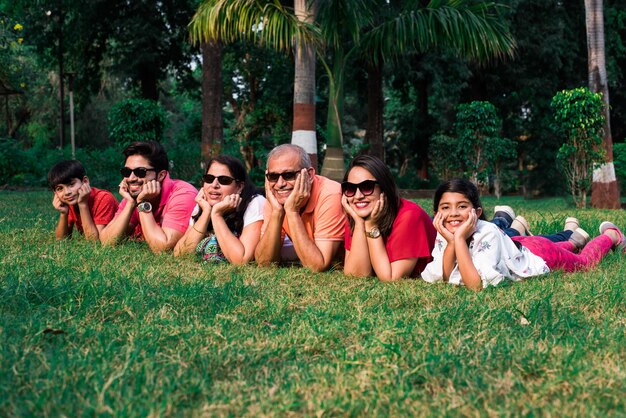 Belle famille indienne multi génération de six personnes, regardant la caméra et souriant en position couchée sur l'herbe verte au jardin ayant les mains sur le menton