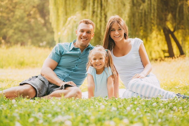 Belle famille souriante assise sur l'herbe dans le parc et regardant la caméra.