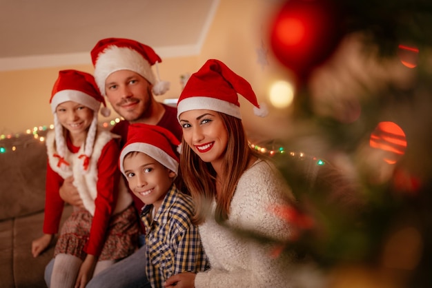 Une belle famille souriante est réunie par un sapin de Noël à la maison. Regarder la caméra et porter des chapeaux de Père Noël.