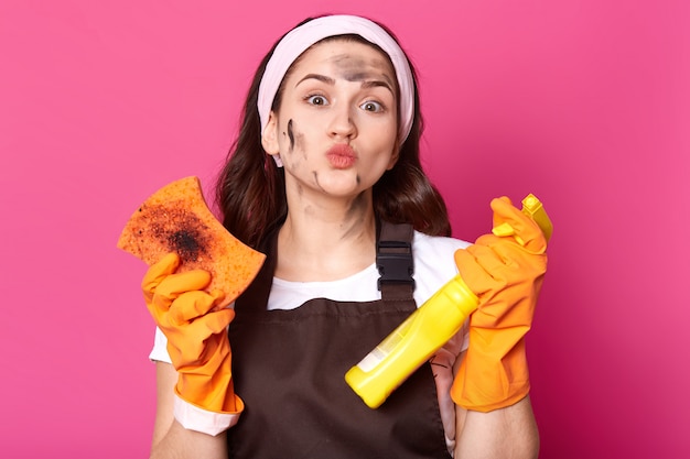 Belle femme adorable avec de la saleté sur son visage qui dépasse ses lèvres, tenant une bouteille de liquide de nettoyage