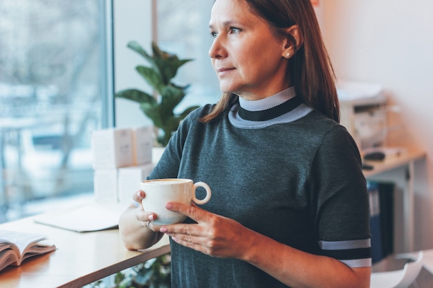 Belle femme adulte d&#39;âge mûr regardant la fenêtre et buvant