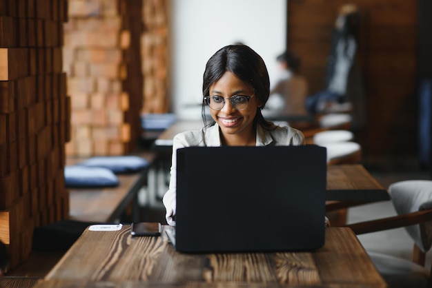 Belle femme d'affaires africaine attendant la réunion dans un espace de coworking et assise avec l'ordinateur portable Hipster girl surfe sur le web sur un ordinateur portable pendant le déjeuner