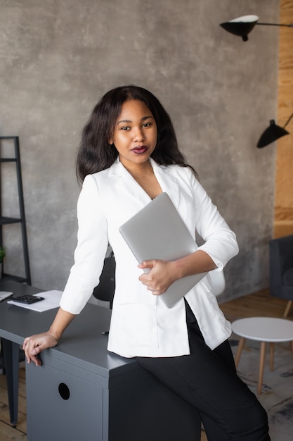 Belle femme d'affaires afro-américaine en costume classique utilise un ordinateur portable et sourit tout en travaillant au bureau