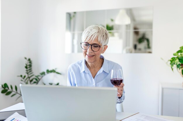 Belle femme d'affaires d'âge moyen portant des lunettes rectangulaires, assise au bureau à domicile avec un verre de vin sur le bureau. Femme aînée travaillant à domicile