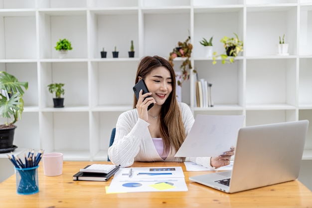 Belle femme d'affaires asiatique assise dans sa chambre, parlant à son partenaire par téléphone portable et vérifiant les documents financiers, elle est une femme cadre d'une start-up. Direction financière.