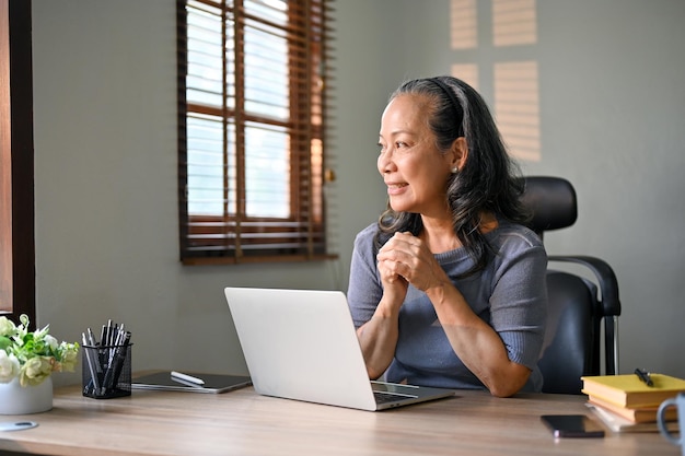 Belle femme d'affaires asiatique est assise à son bureau et regarde par la fenêtre en rêvant