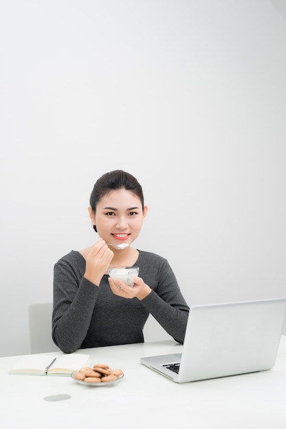 Belle femme d'affaires asiatique mange du yaourt et sourit en étant assise sur son lieu de travail à la maison