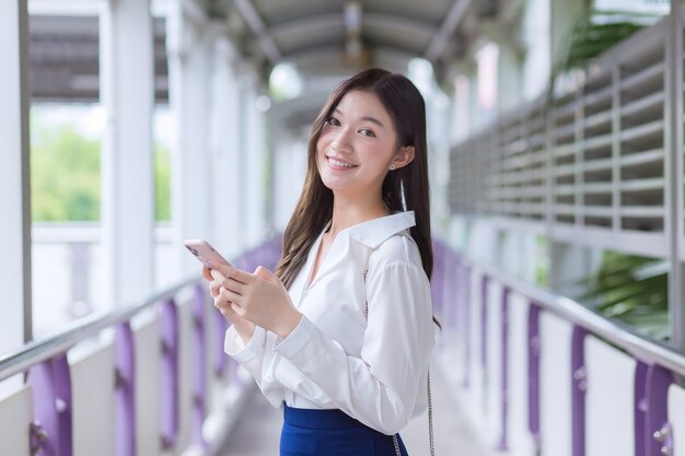 Une belle femme d'affaires asiatique se tient sur le survol du train aérien en ville tout en utilisant son smartphone