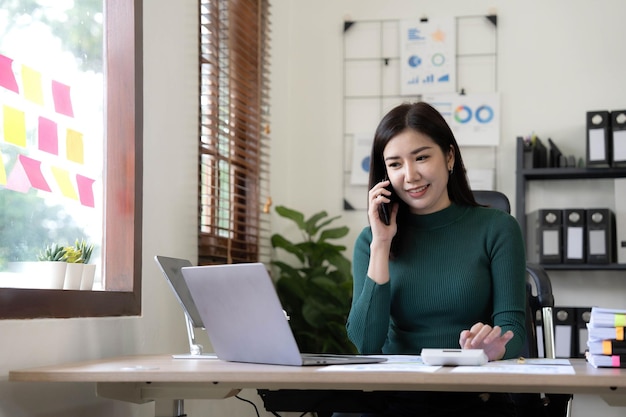 Belle femme d'affaires asiatique souriante analysant un tableau et un graphique montrant les changements sur le marché et tenant un smartphone au bureau