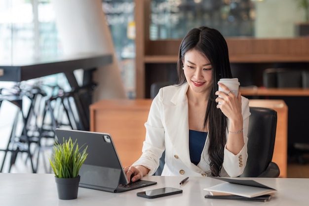 Belle femme d'affaires asiatique tenant un café assis sur une chaise de bureau à l'aide d'un clavier de tablette.