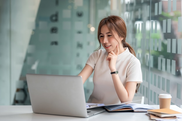 Belle femme d'affaires asiatique travaillant sur un ordinateur portable et des documents dans un bureau moderne.