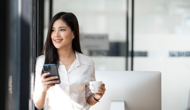 Une belle femme d'affaires asiatique utilisant un téléphone portable alors qu'elle se tient au bureau en levant les yeux et en tenant une tasse de café