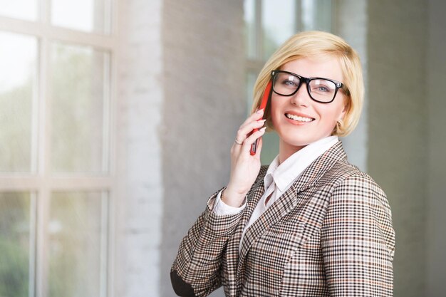 Belle femme d'affaires blonde vêtue d'un costume élégant à carreaux avec des lunettes parlant avec un smartphone