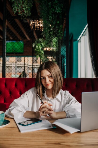 Une belle femme d'affaires brune en chemise blanche est assise à une table dans un café avec un ordinateur portable