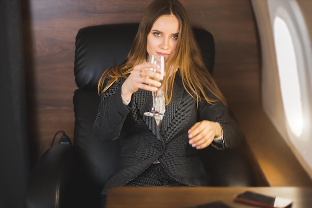 Une belle femme d'affaires caucasienne confiante en costume, maquillée, cheveux longs assise dans un jet privé avec une coupe de champagne, célébrant le succès. Concept d'entreprise.