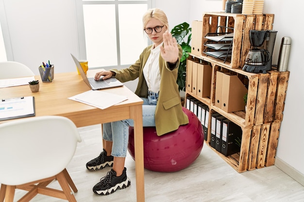 Belle femme d'affaires caucasienne travaillant au bureau assise sur une balle de pilates avec la main ouverte faisant un panneau d'arrêt avec une expression sérieuse et confiante, geste de défense